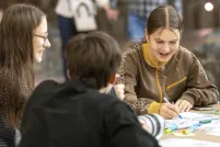 Two teenage girls sit at a table, laughing and colouring in a design with markers, while another person sits nearby with their back to the camera.
