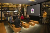 A small group of people sitting on wooden benches in a theatre face a screen showing an older woman seated in a chair. A man in a yellow shirt is standing to the right of the screen holding a microphone.
