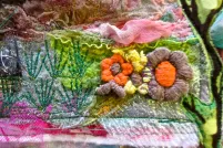 A colourful textile art piece that includes embroidered flowers, branches and greenery mixed with layers of pink, blue and white fabrics. 
