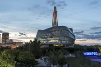 Exterior photo of the Canadian Museum for Human Rights’ glass windows and tower, taken against a prairie sky at dusk. The Winnipeg sign at the Forks is situated on the right side of the image. Multiple office towers and downtown lights are on the left-hand side of image.