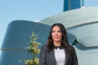 A woman with long dark hair, wearing a suit jacket, stands with her hands clasped in front of her. Behind her is a large, unusual building with a rounded glass façade and a tower.