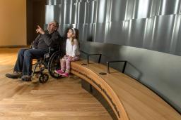 An older man sits in a wheelchair beside a young girl sitting on a bench. The man is pointing up, and both he and the young girl are looking up and to the viewer’s left.
