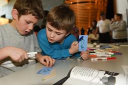 Two young boys holding markers and small cards with string and reading one of the cards.