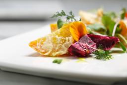 Bright gold-coloured potato wedges are artfully arranged on a square white plate, along with leaf greens, wedges of red beets, and garnish.