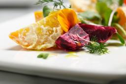 Bright gold-coloured potato wedges are artfully arranged on a square white plate, along with leaf greens, wedges of red beets, and garnish.