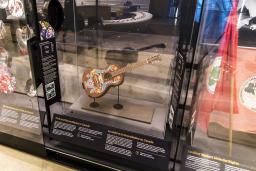 A row of museum display cases with various artifacts inside. A guitar is displayed under glass. The guitar has a colourful floral print with a bird hand-painted onto its front.