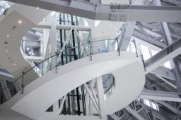 A view in clear daylight looking up at an elevator shaft and a staircase that winds around it. The framework of the elevator shaft is visible. Steel beams can be seen all around the shaft and staircase.