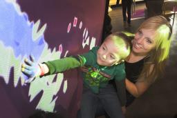 A woman and a young boy look up at a wall. The boy is reaching out and touching the wall. Blue, red and green coloured light is being projected onto the wall and the boy’s hand.