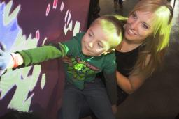 A woman and a young boy look up at a wall. The boy is reaching out and touching the wall. Blue, red and green coloured light is being projected onto the wall and the boy’s hand.