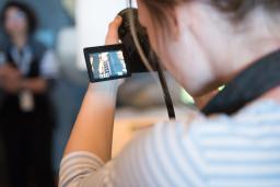 A person looks through the screen of a camera in the foreground while a person poses in the background.