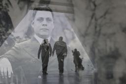 A translucent historical image of a man in a suit layered over a contemporary photo showing the dark outline of two men walking in a triangular hallway.