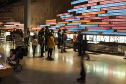 A large number of people in a gallery with a wall of colourful panels and photos showing a timeline of events.