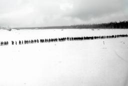 A black-and-white photo of a flat, snowy landscape with trees and small buildings in the distance. A long, straight line of more than 100 people are walking through snow.