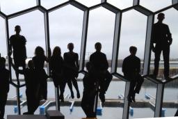 Eighteen people in a row, some seated, some standing, in front of a wall of odd-shaped windows. 