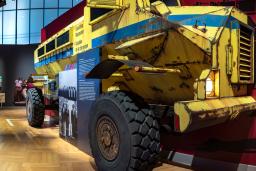 A museum gallery with large, yellow armoured truck emerging from one wall and rusty garbage-can lids hanging on the opposite wall.