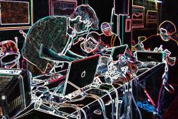 Four people around a table covered in computers and electronic music equipment. The photo has been altered so that the background is black and the elements of the photo are outlined in multi-coloured neon.