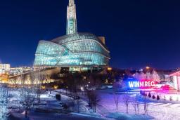 A glass and steel building on a snowy evening, positioned behind an illuminated sign that says Winnipeg.