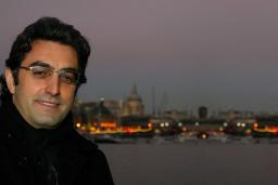 Close-up of a smiling man at dusk. Behind him is a body of water and on the other side there are boats, lights and buildings. 