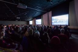 A seated audience viewing three large screens in a dimly lit room.