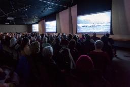 A seated audience viewing three large screens in a dimly lit room.