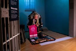 An Indigenous man wearing ceremonial regalia kneels inside a replica of a prison cell. He is presenting a textile banner with a graphic image of a bird and the words “bring our children home.” 