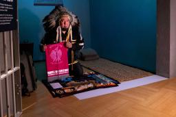 An Indigenous man wearing ceremonial regalia kneels inside a replica of a prison cell. He is presenting a textile banner with a graphic image of a bird and the words “bring our children home.” 