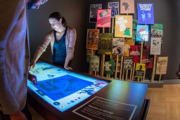 A man and woman look down at an illuminated table. In the background, there is a grouping of colourful posters on the wall.