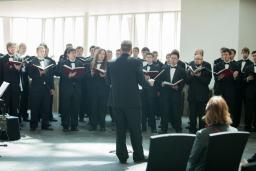 A choir of men wearing black outfits hold books and sing in front of a conductor.