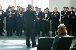 A choir of men wearing black outfits hold books and sing in front of a conductor.