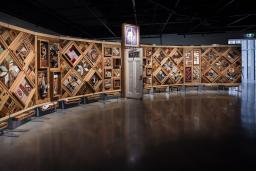 A large horizontal art installation with a wooden frame made up of multiple diamonds and rectangles to which many objects are mounted. There is a half-opened door in the middle of the work.