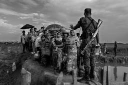A soldier with a rifle on his back blocks a group of Rohingya before they cross a makeshift bridge. His left hand is raised, gesturing for them to stop.
