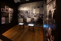 A Museum exhibit showing a black-and-white photo of children sitting in rows at school desks. Two desks, similar to those in the photo sit in the centre of the exhibit. A headline on a text panel reads “Childhood Denied.”