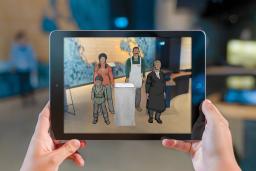 A visitor's hands hold a tablet that carries an image of four animated people including a little boy, a young Indigenous woman, a man wearing an apron and a woman judge.