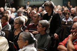 A room full of audience members sitting. One woman is standing and smiling holding a microphone.