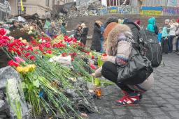 Two people laying red flowers at a memorial. In the background a dozen other people are standing in front of debris, such as rocks, metal sheeting and cement barriers painted with blue and yellow Ukrainian flags and lettering. 