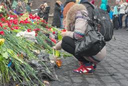 Two people laying red flowers at a memorial. In the background a dozen other people are standing in front of debris, such as rocks, metal sheeting and cement barriers painted with blue and yellow Ukrainian flags and lettering. 