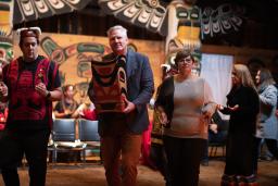 Two men and a woman dance side by side. One man is wearing a red vest and a headdress. The other man, in a suit jacket, carries a bentwood box carved in west coast Indigenous style. In the background are two larger totem poles and people dressed in regalia.