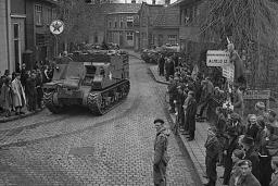 A crowd gathered on sidewalks, watching armoured vehicles driving through the cobblestone streets of a village.