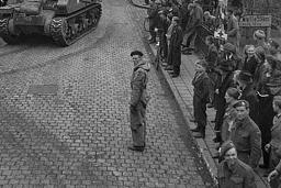 A crowd gathered on sidewalks, watching armoured vehicles driving through the cobblestone streets of a village.