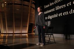 A young woman talks in front of a black wall that says, in large white letters, “Tous les êtres humains naissent libres et égaux en dignité et en droits.” (All human beings are born free and equal in dignity and rights.) There is a circular theatre in the background that resembles a large woven basket.