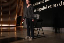 A young woman talks in front of a black wall that says, in large white letters, “Tous les êtres humains naissent libres et égaux en dignité et en droits.” (All human beings are born free and equal in dignity and rights.) There is a circular theatre in the background that resembles a large woven basket.