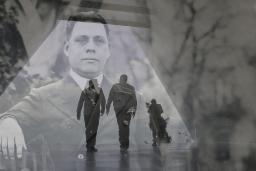 A translucent historical image of a man in a suit layered over a contemporary photo showing the dark outline of two men walking in a hallway.