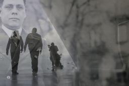 A translucent historical image of a man in a suit layered over a contemporary photo showing the dark outline of two men walking in a hallway.