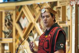 A man wearing traditional Indigenous regalia and headdress stands in front of an art installation made of wooden panels with embedded objects.
