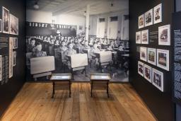 A museum exhibit that includes small school desks in front of a large photographic background of Indigenous children sitting at their desks in a classroom.