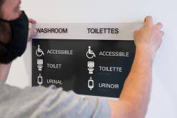 A man holds up a sign that says "Washroom" as it is mounted to a wall. Underneath the title, the words "accessible," "toilet" and "urinal" are accompanied by descriptive icons.