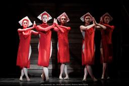 On a dark stage are five ballet dancers dressed in red knee-length flowy costumes with white square-like hats. They are looking forward with their hands in prayer.