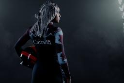 A black woman stands in a dark place between two large projectors. She's holding a helmet and wears a Canadian bobsleigh suit.