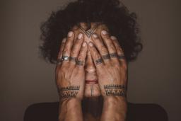 Aaju Peter covers her eyes and cheeks with her bare hands. Tattoos are visible on her forehead, chin, fingers and wrists. Her hands and the visible parts of her face are illuminated and are framed by her dark curly hair and angular shoulders.