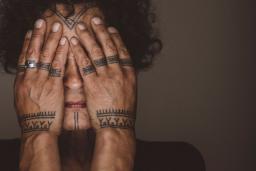 Aaju Peter covers her eyes and cheeks with her bare hands. Tattoos are visible on her forehead, chin, fingers and wrists. Her hands and the visible parts of her face are illuminated and are framed by her dark curly hair and angular shoulders.